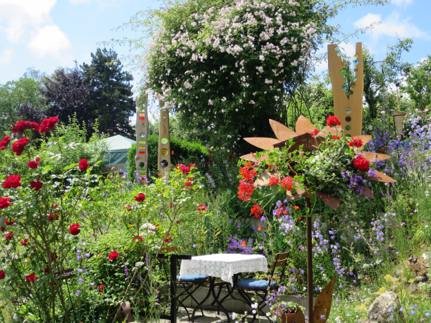Ein Garten mit bunten Blumen und Pflanzen, der einen zentralen Tisch mit einer Decke und Stühlen zeigt, umgeben von Bäumen unter einem bewölkten Himmel auf der Chelsea Flower Show.