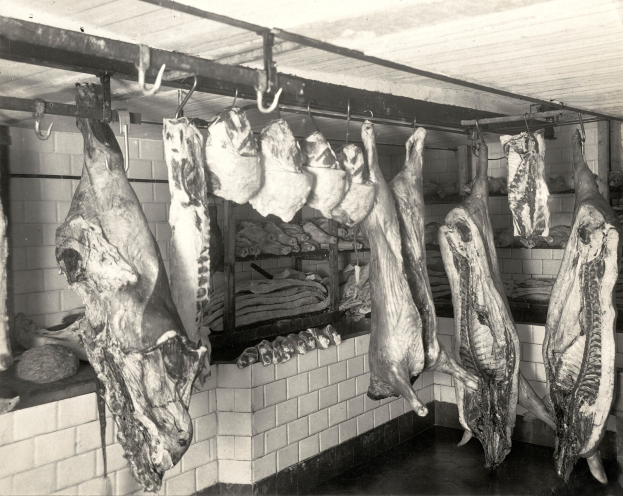 Ein Schwarz-Weiß-Foto einer Metzgerei mit verschiedenen Fleischstücken, die von der Decke hängen, einer Wand im Hintergrund und einer Decke oben.