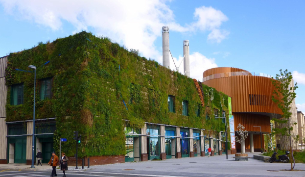Großes nachhaltiges Gebäude mit einer grünen Wand, umgeben von Straßeninfrastruktur, Fahrzeugen, Fußgängern, Bäumen und einer wolkigen Himmel.