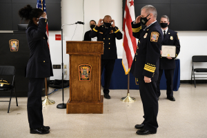 Gruppe uniformierter Personen in Masken vor einem Podium mit Mikrofon, umgeben von Fahnen und Möbeln, bei einer Vereidigung, bei der eine Person ein Zertifikat hält.