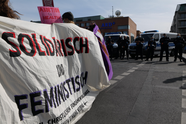 Eine Gruppe von Menschen marschiert auf einer Straße, hält ein "Solidarität und Feminismus"-Schild, mit geparkten Fahrzeugen, Gebäuden, einer Schüsselantenne und einem klaren blauen Himmel im Hintergrund.