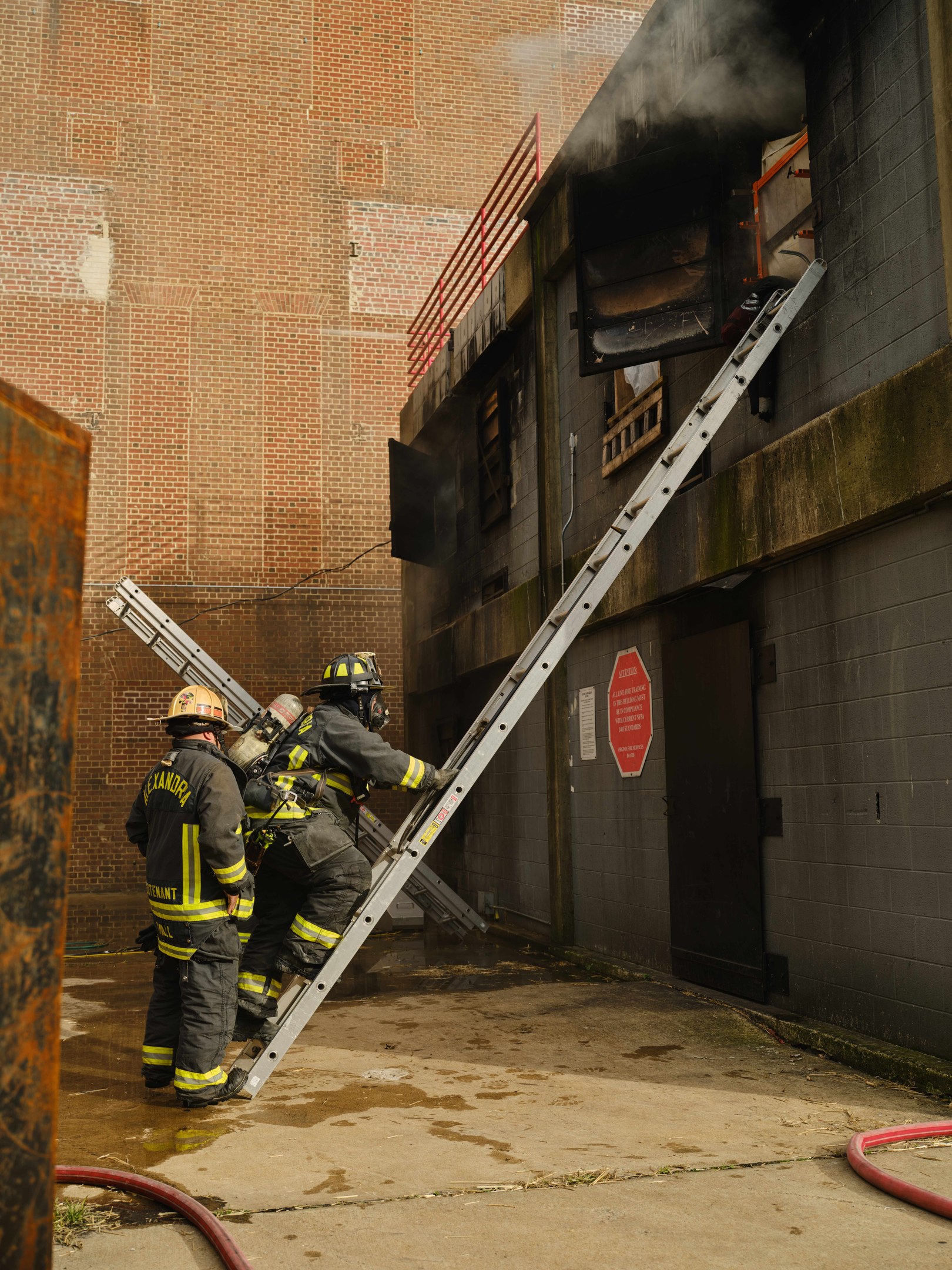Feuerwehrleute in Helmen klettern eine Leiter hinauf zu einem rauchgefüllten Gebäude, mit einem Metallobjekt links und Rohren am Boden.