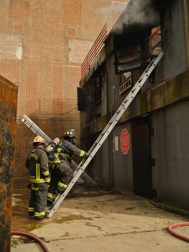 Feuerwehrleute in Helmen klettern eine Leiter hinauf zu einem rauchgefüllten Gebäude, mit einem Metallobjekt links und Rohren am Boden.