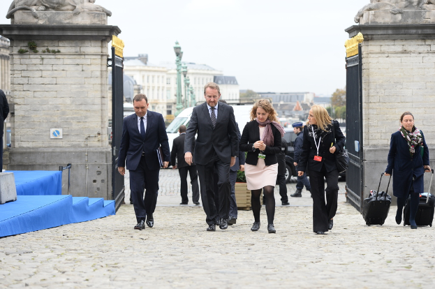 Eine Gruppe von Mitarbeitern geht in einen Bereich mit einem Tor, hinter dem sich viele Fahrzeuge und Gebäude befinden, während eine Frau rechts zwei Gepäckstücke trägt.