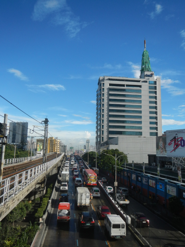 Eine belebte Stadtstraße mit starkem Verkehr, hohen Gebäuden, einer Brücke auf der linken Seite, Strommasten mit Drähten, Bäumen und einem bewölkten Himmel im Hintergrund.