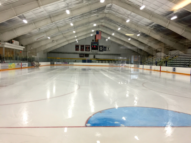 Große Indoor-Eisfläche mit Deckenbeleuchtung, Werbetafeln, einer Wand, Treppen mit Geländern und einigen Menschen, wahrscheinlich eine Hockey-Einrichtung.