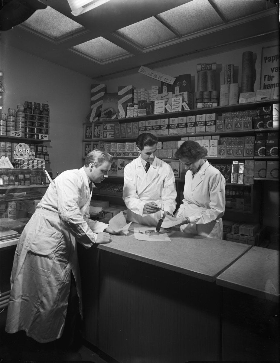 Drei Männer in weißen Kitteln an einer Apotheken-Theke mit Papieren und Gegenständen, vor Regalen mit Artikeln, in Schwarz-Weiß.