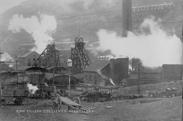 Schwarz-weiß-Foto einer rauchenden Fabrik umgeben von Gebäuden, Pfählen, Fahrzeugen, Karren und anderen Gegenständen mit Text unten.