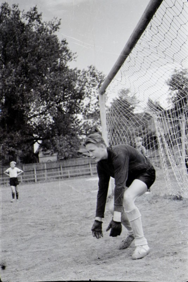Schwarz-weiß-Foto eines jungen Jungen, der auf einem Feld mit einem Zaun, Bäumen und einem klaren Himmel Fußball spielt, mit einer Person im Vordergrund und anderen Kindern im Hintergrund.