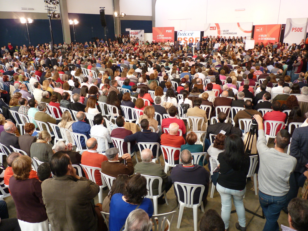 Ein volles Stadion mit sitzenden und stehenden Menschen, eine Bühne mit zwei Rednern an einem Podium und nahegelegenen Bannern und Lichtern.