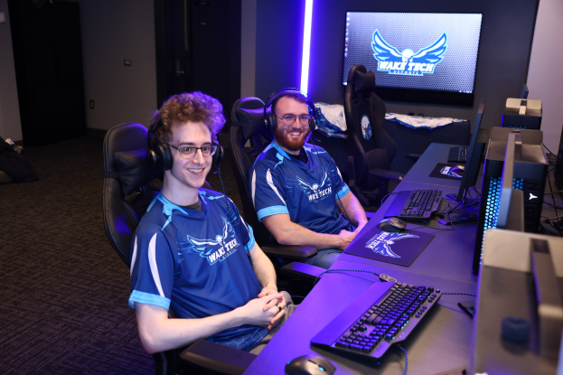 Zwei Männer mit Headsets sitzen an einem Schreibtisch mit Monitoren, Tastaturen und anderer Ausrüstung und lächeln, während sie ein Spiel der Wake Tech League of Legends zu spielen scheinen.