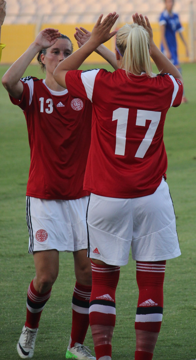 Zwei Frauen in roten T-Shirts und weißen Shorts klatschen auf einem Fußballfeld mit einer Person in einem blauen Kleid, einer gelben Tafel und Stühlen im Hintergrund.