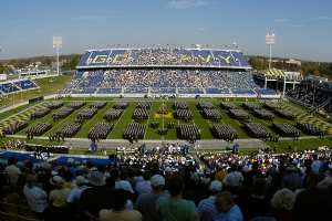 Eine große Menschenmenge in einem Stadion, die ein Fußballspiel anguckt, mit einigen Stehenden und anderen Sitzenden auf einem Rasenfeld unter einem klaren blauen Himmel, mit Laternenmasten, Bäumen und Gebäuden im Hintergrund.