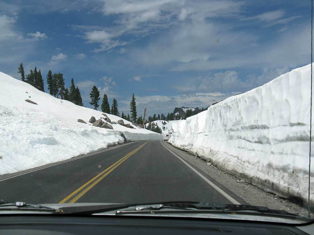 Ein Blick aus einem Fahrzeug, der eine leere Straße mit schneebedeckten Bergen und Bäumen am Straßenrand zeigt.