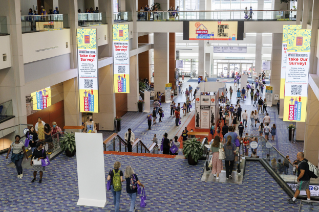 Eine große Menschenmenge durchquert ein großes Gebäude mit Bannern, Topfpflanzen, Rolltreppen, Geländern, Säulen, bedruckten Tafeln, Glastüren und anderen Elementen, einige tragen Taschen, was auf den Besuch der Nationalen Buchmesse hindeutet.