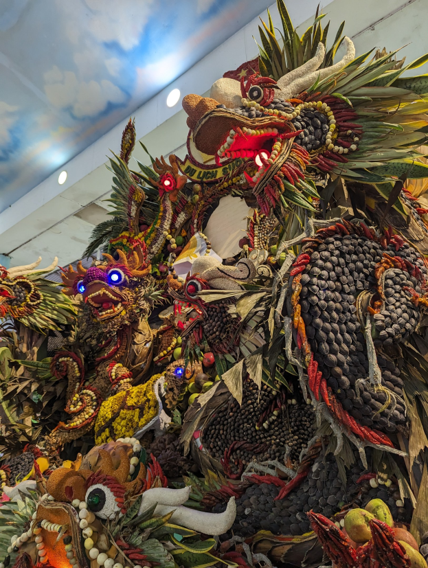 Ein großer Haufen Obst und Gemüse in einem Raum mit beleuchteter Decke und einer Wand im Hintergrund, mit dem Text 'Papua-Neuguinea-Karnival' unten.