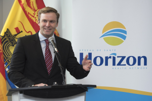 Ein l├Ąchelnder Mann in einem schwarzen Blazer, wei├čem Hemd und roter Krawatte steht an einem Podium mit einem Mikrofon und spricht als Gesch├Ąftsführer mit einem Banner im Hintergrund.