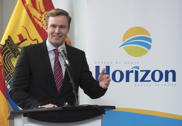 Ein l├Ąchelnder Mann in einem schwarzen Blazer, wei├čem Hemd und roter Krawatte steht an einem Podium mit einem Mikrofon und spricht als Gesch├Ąftsführer mit einem Banner im Hintergrund.