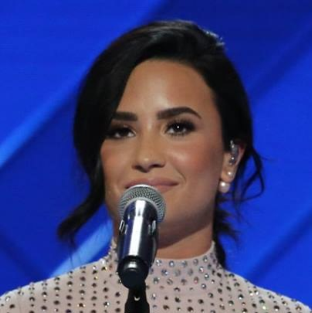Demi Lovato steht selbstbewusst bei den Grammys mit einem eleganten Bob, trägt ein schwarzes Kleid und steht vor einem Mikrofon vor einem blauen Hintergrund.