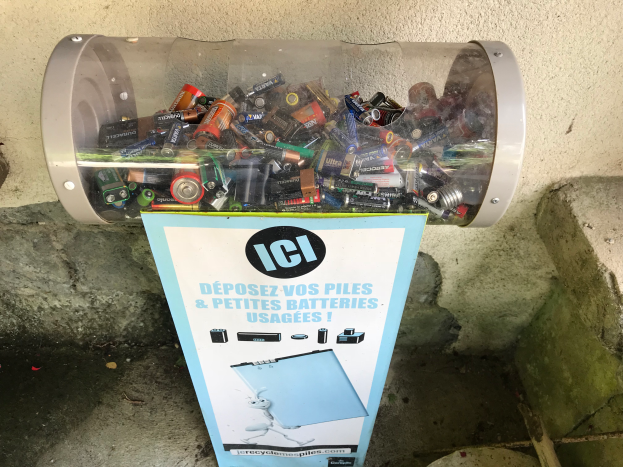 Ein Müllcontainer voller Batterien auf einem Bürgersteig, mit einem Plakat mit Text an der Wand dahinter und einer Treppe auf der rechten Seite.