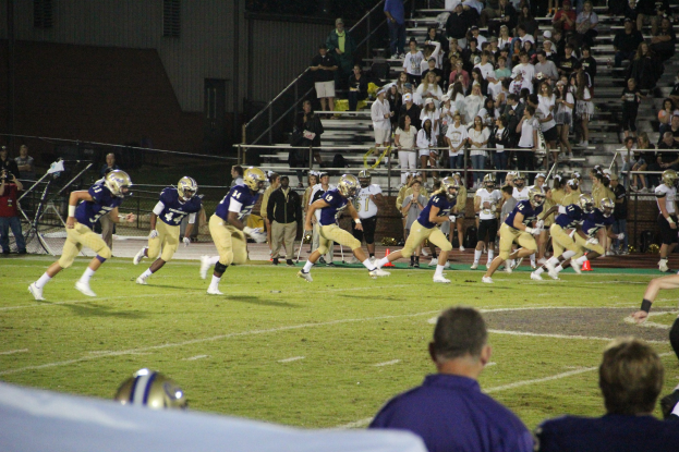 Ein Fussballspiel wird auf einem Rasenfeld mit Spielern in Sportkleidung und Helmen im Vordergrund gespielt, während Zuschauer auf Treppen sitzen und im Hintergrund an Geländern und einem Gebäude stehen, mit einer Texttafel am unteren Bildrand.