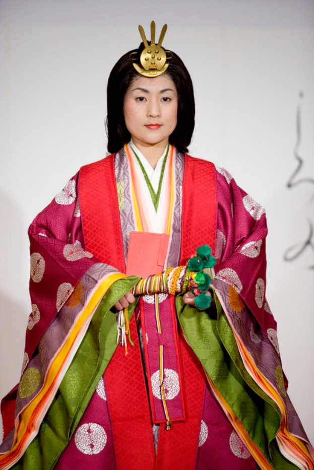 Eine Frau in einem traditionellen japanischen Kimono, das ein leuchtendes rotes und grünes Kleid mit aufwändigen Mustern trägt, steht selbstbewusst auf einem Laufsteg, ihr Haar ist ordentlich zu einem Knoten gebunden.