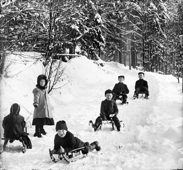 Eine Gruppe von Kindern auf Rodelschlitten, die einen schneebedeckten Hügel mit Bäumen im Hintergrund hinunterrutschen, dargestellt in Schwarz-Weiß.