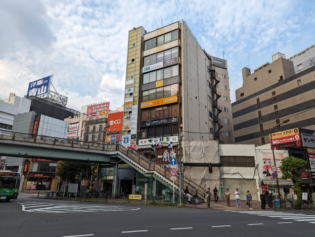Eine belebte Stadtstraße mit einem Bus, hohen Gebäuden, Schildern, einer Brücke, Fußgängern, Fahrzeugen, einem Zaun, Bäumen, Straßenlaternen und einer bewölkten Himmel.