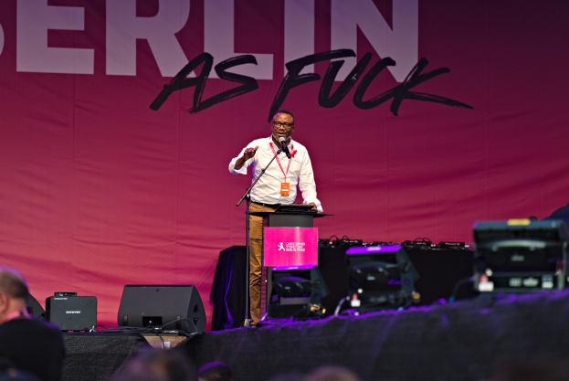 Mann in weißem Hemd und braunen Hosen steht auf einer Bühne und hält ein Mikrofon, während er eine Menge hinter einem pinken Banner mit der Aufschrift 'Berlin as Fuck' anspricht.