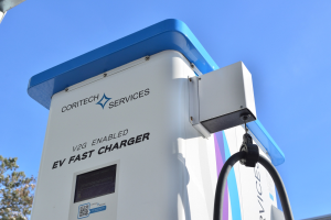 Elektrische Fahrzeugladestation mit einem blauen und weißen "V2G-Fast-Charger"-Schild, Bäumen, einem Gebäude und einem klaren blauen Himmel.