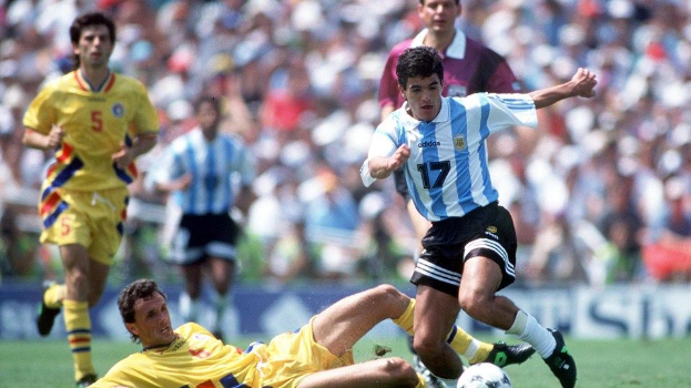 Eine Gruppe von Männern, die auf einem grünen Feld Fußball spielen, mit Argentiniens Diego Maradona, der von Kolumbiens Diego Sanchez getackelt wird, umgeben von Zuschauern und Tafeln.