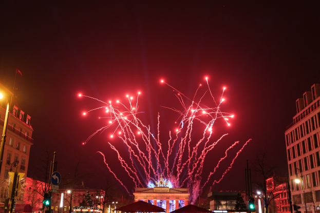 Eine Straßen in Berlin an Silvester, voller Gebäude, Bäume, Laternenpfähle, Verkehrszeichen, Zelte und Menschen, mit einem Feuerwerk erleuchteten Himmel im Hintergrund.