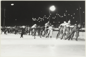 Eine Gruppe von Menschen, die nachts auf einer Eisbahn Schlittschuh laufen, beleuchtet von Straßenlaternen im Hintergrund, auf einem Schwarz-Weiß-Foto.