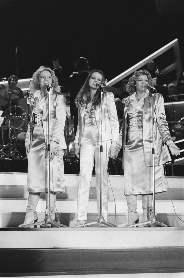 Drei Frauen singen in Mikrofone auf der Bühne mit Musikern im Hintergrund, schwarz-weiß.