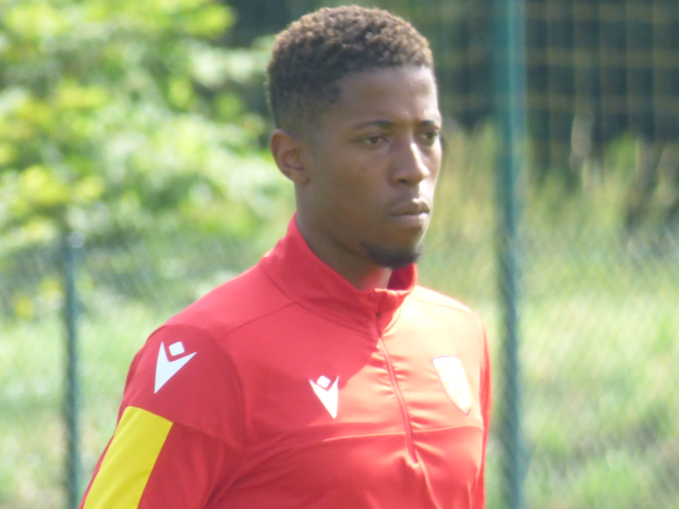 Ein junger Mann in einem roten und gelben T-Shirt steht auf einem Fußballfeld, das von einem Zaun und Bäumen umgeben ist und sieht entschlossen aus.