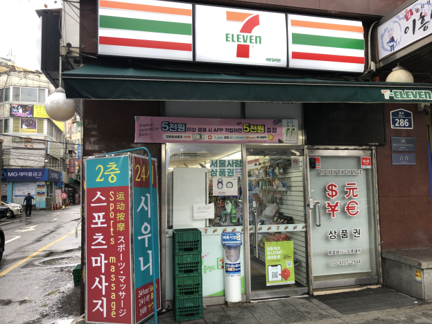 Ein 7-Eleven-Konzern in Seoul, Südkorea, mit Fahrzeugen auf der Straße, einem Fußgänger auf dem Gehweg und Gebäuden im Hintergrund unter einem sichtbaren Himmel.