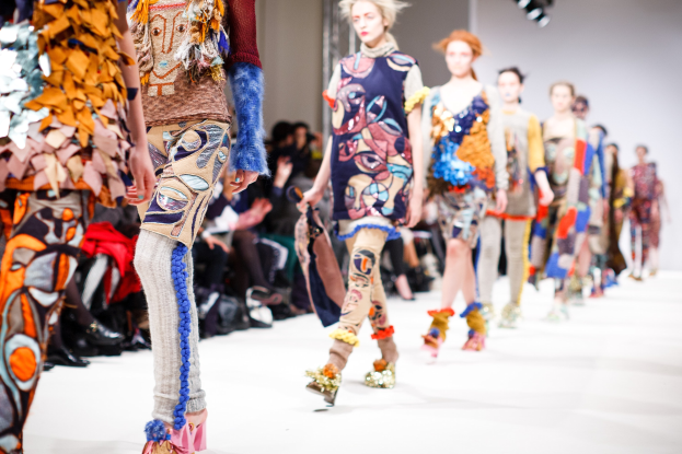 Eine Gruppe Von Frauen, Die Eine Laufsteg In London Fashion Week Heruntergehen, Mit Einer Zuschauerschaft Im Hintergrund Unter Helligen Lichtern.