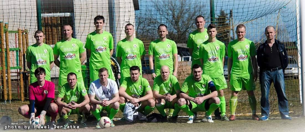 Gruppe von Männern in grünen T-Shirts auf einem Fußballfeld neben einem Torpfosten mit einem Ball auf dem Boden und verschiedenen Hintergrundelementen.