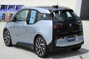 Ein BMW i3 Elektroauto vor einem Gebäude geparkt, mit einer Pflanze im Vordergrund und einer Wand im Hintergrund; das Auto hat ein Rad in der Mitte des Bildes.