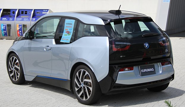 Ein BMW i3 Elektroauto vor einem Gebäude geparkt, mit einer Pflanze im Vordergrund und einer Wand im Hintergrund; das Auto hat ein Rad in der Mitte des Bildes.
