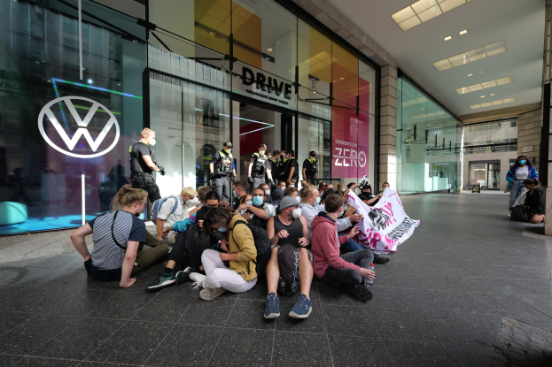 Eine Gruppe von Menschen sitzt auf dem Boden vor einer Volkswagen-Vertretung mit Glaswänden, die eine Fahne mit Text halten, unter Deckenleuchten, die zu protestieren scheinen.