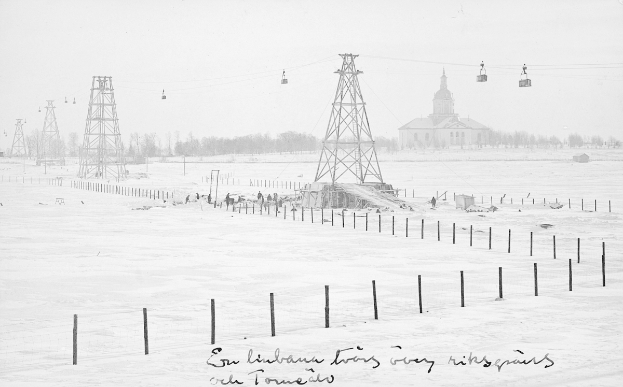 Ein Schwarz-Weiß-Foto einer Skiliftanlage in einer verschneiten Landschaft, umgeben von Pfählen, Seilen, Bäumen und einem Gebäude im Hintergrund, mit Text am unteren Bildrand.
