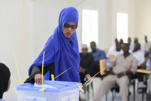 Eine Frau mit einem blauen Kopftuch stimmt an einer Wahllokal ab, während andere in der Nähe sitzen.