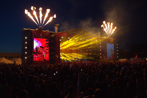 Eine große Menschenmenge auf einem Musikfestival, mit einer Bühne in der Mitte, auf der Musiker Instrumente spielen, beleuchtet von hellen Lichtern und umgeben von Bannern, Zelten links und einem dunklen Himmel mit Feuerwerk im Hintergrund.