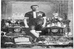 Schwarzes und weißes Foto eines Mannes in einem Fußballtrikot, der vor einem Tisch mit Trophäen und Pokalen steht, mit der Beschriftung "1909-1910 WM-Sieger" am unteren Rand.