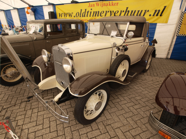 Vintage-Roadster vor einem blauen und weißen Zelt mit einer Fahne und Luftballons im Hintergrund.