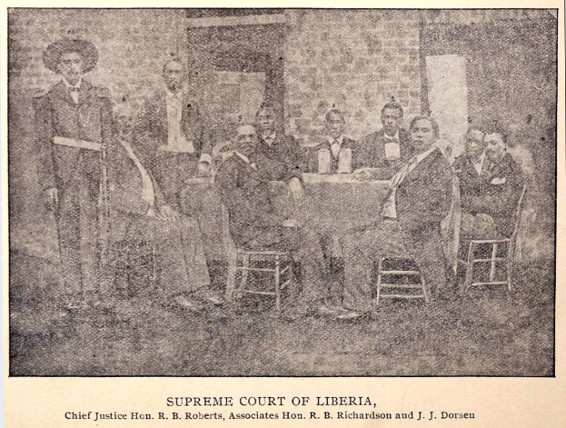 Eine Gruppe von Männern, die um einen Tisch vor dem Obersten Gerichtshof von Liberia sitzen, mit einer Wand und einer Tür im Hintergrund.