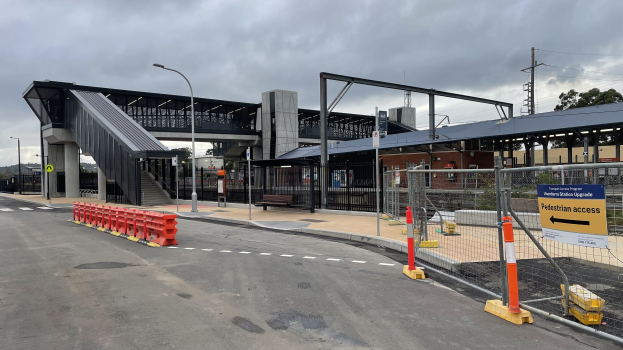 Ein Bahnsteig mit einer Brücke im Hintergrund, Straßenlaternen, Strommasten, Stromkabel, Bäume, Gebäude, Fahrzeuge auf der Straße, Absperrpoller, Schilder, Bänke und eine bewölkte Himmel.