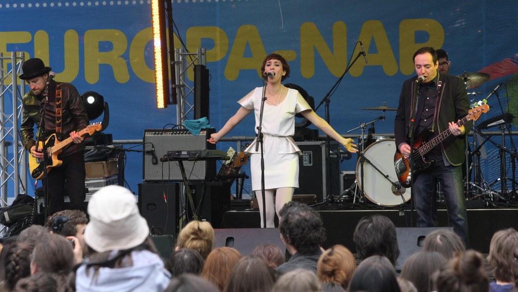 Vier Musiker auf der Bühne: eine Frau in einem weißen Kleid singt in der Mitte, flankiert von zwei Männern mit Gitarren, mit Publikum darunter und Equipment im Hintergrund.