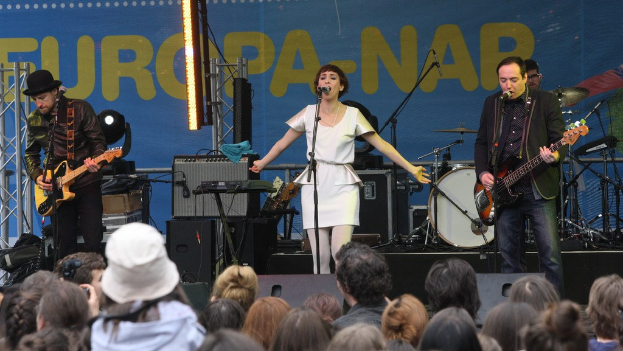 Vier Musiker auf der Bühne: eine Frau in einem weißen Kleid singt in der Mitte, flankiert von zwei Männern mit Gitarren, mit Publikum darunter und Equipment im Hintergrund.